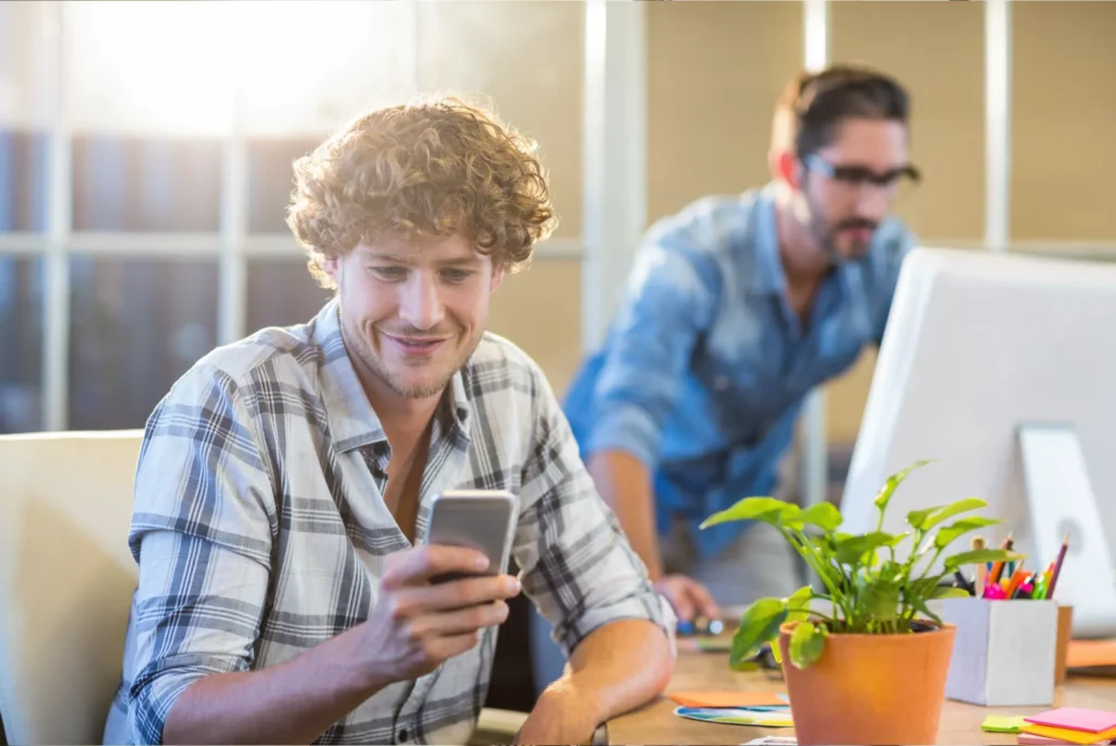 Hell erleuchtetes Büro mit zwei jungen Männern, einer schaut auf sein Smartphone, Pflanzen auf dem Tisch; Szene in einer Agenturumgebung bei Luxregia