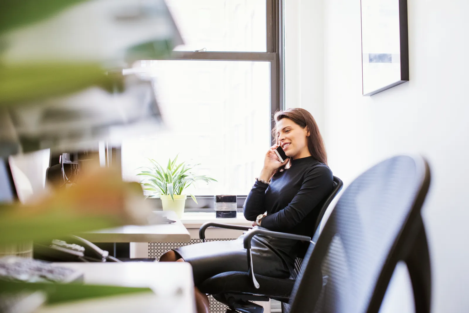 Eine Frau in einem hellen Büro sitzt an einem Schreibtisch, spricht am Smartphone, trägt ein schwarzes Oberteil; im Hintergrund ein Fenster mit Tageslicht, eine grüne Pflanze auf dem Tisch.