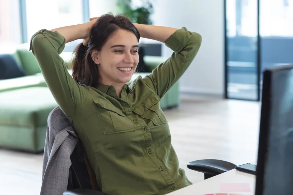 Eine caucasian Frau in grünem Hemd lehnt sich entspannt zurück und sitzt am Schreibtisch in einem hellen Büro, im Hintergrund eine moderne Arbeitsatmosphäre – Kontext: Luxregia-Prozess-Automatisierung