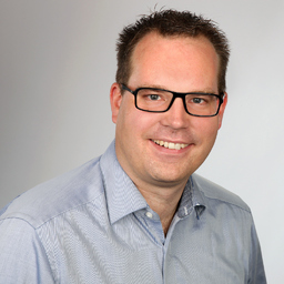 Christopher Fabry mit schwarzer Brille und hellblauem Hemd vor hellem Hintergrund, Portraitfoto eines Mitarbeiters von Luxregia.