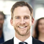 Lächelnder Joachim Ficht im Anzug mit gepflegtem Bart und kurzen braunen Haaren, klarer Blick, helles Licht im Hintergrund.
