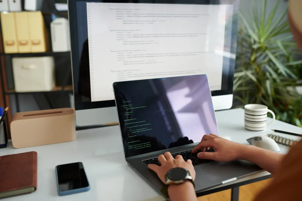 Luxregia GmbH Arbeitsumgebung: Entwickler tippt Code am Laptop, Monitor im Hintergrund zeigt Programmierzeilen, moderne Büroausstattung mit Smartphone und Notizbuch