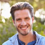 Lächelnder junger Mann mit verschränkten Armen, freundlicher Gesichtsausdruck, helles Oberteil, weicher Hintergrund im Freien—stimmige natürliche Stimmung.