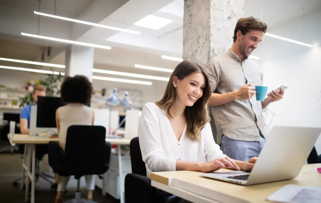 Hell beleuchtete Bürofläche mit zwei Mitarbeitenden: Eine Frau tippt am Laptop, während ein Mann im Hintergrund einen Becher hält und ein Smartphone benutzt; Logo der Luxregia GmbH im Kontext einer Marketing-Agentur.