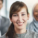 Lächelnde Celina Manka mit braunem Haar und grauem Strickpullover im Büro