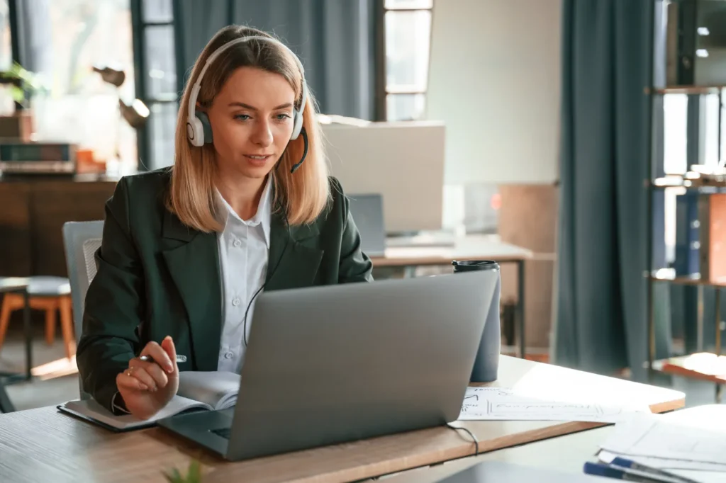 Luxregia GmbH Mitarbeiterin mit Kopfhörern am Laptop im Büro, helle Beleuchtung, formeller Blazer, Unterlagen liegen auf dem Tisch