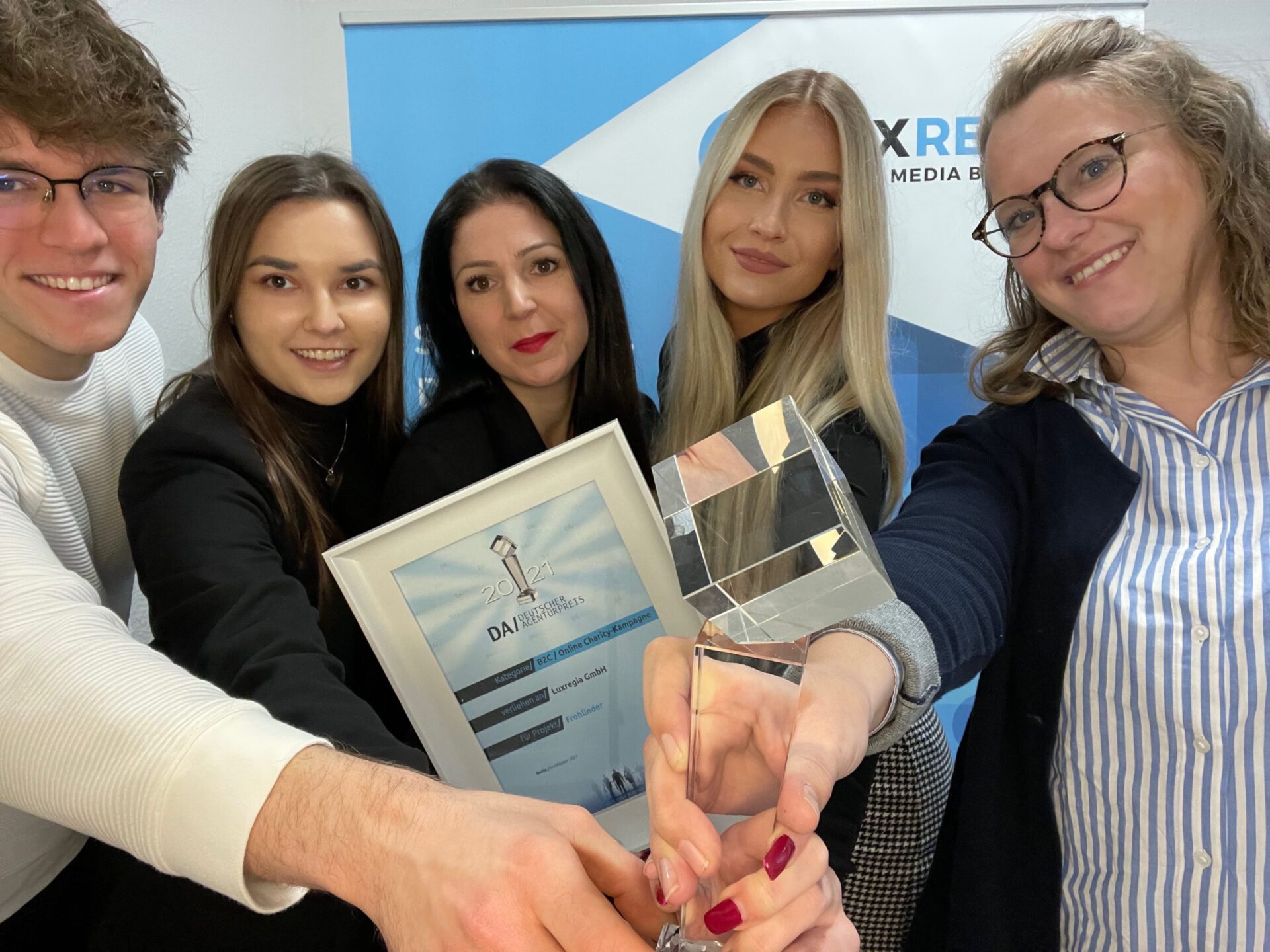 Luxregia Team beim Deutschen Agenturpreis mit Glaspokal und Urkunde vor blauem Messedisplay, vier Personen in Business-Outfits.