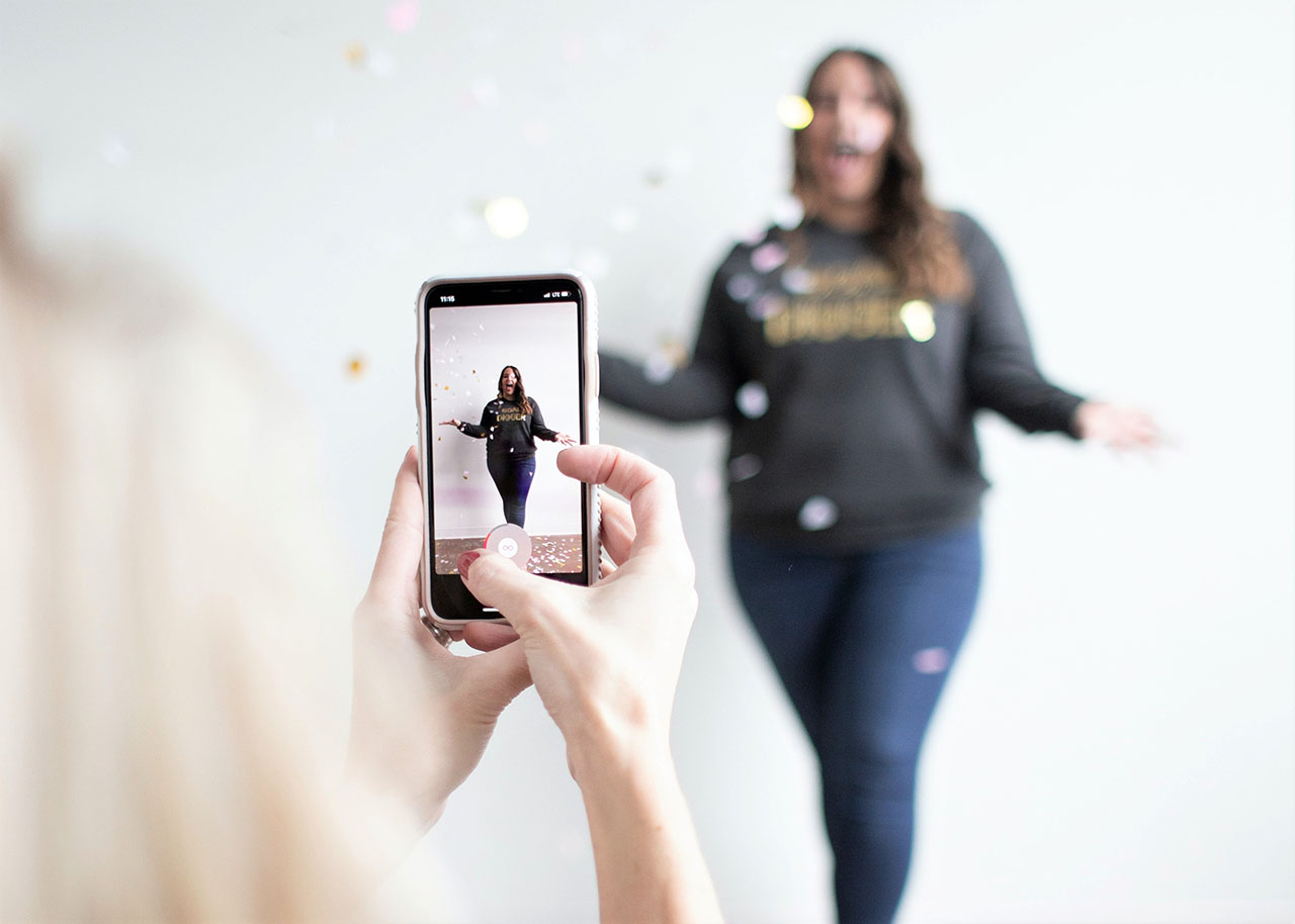 Hand hält Smartphone mit sichtbarem Instagram Reels Video im Hochformat, im Hintergrund eine Person; hell-weißer Look, blaue Kleidung, Facebook-Design