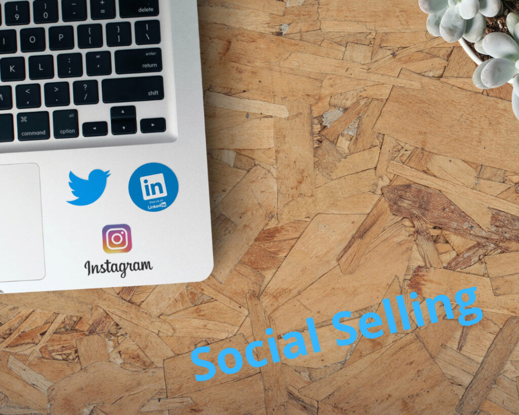 Laptop mit Twitter-, LinkedIn- und Instagram-Icons sowie Aufschrift „social selling“ in Blau auf Holz – Social Selling für Einsteiger.